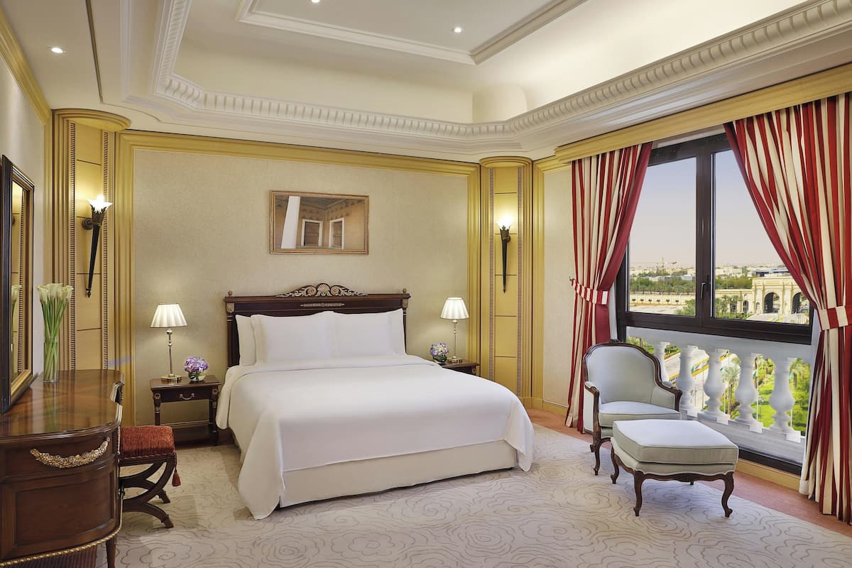 Quarto de hotel com uma cama grande, uma cadeira, uma escrivaninha, um abajur e uma janela com cortinas.
