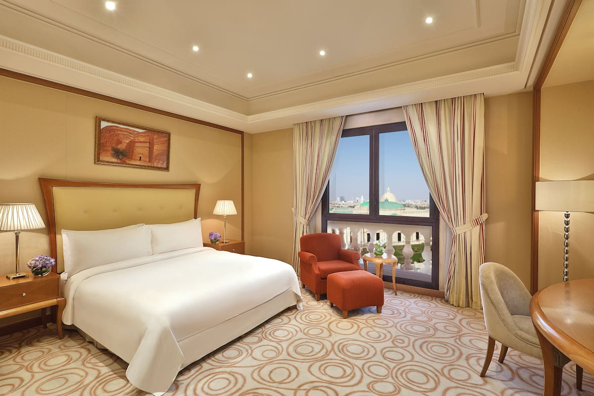 Quarto de hotel com uma cama grande, uma cadeira, uma mesinha e uma janela com cortinas.
