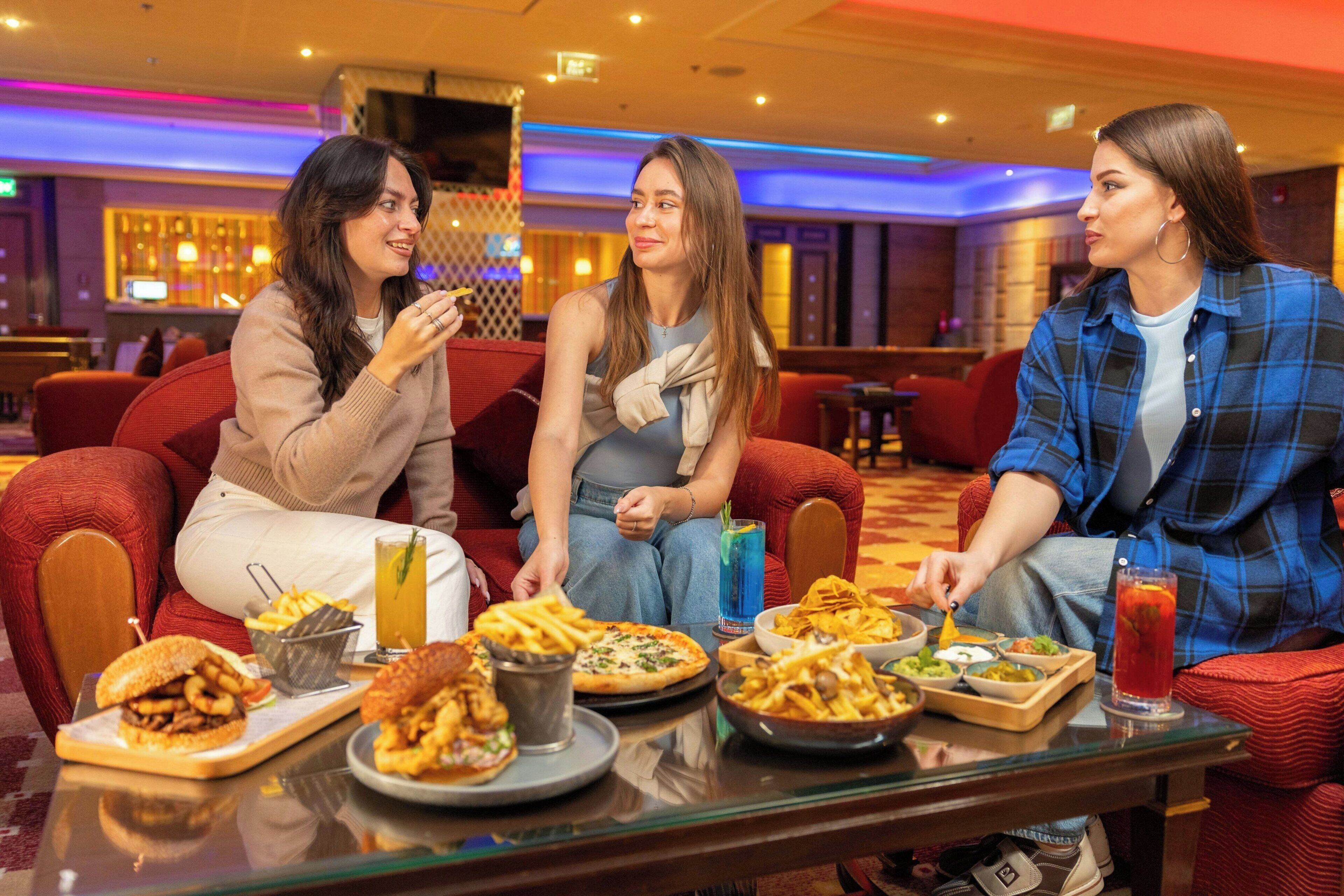 Three people seated on red couches in a restaurant with various dishes and drinks on the table.