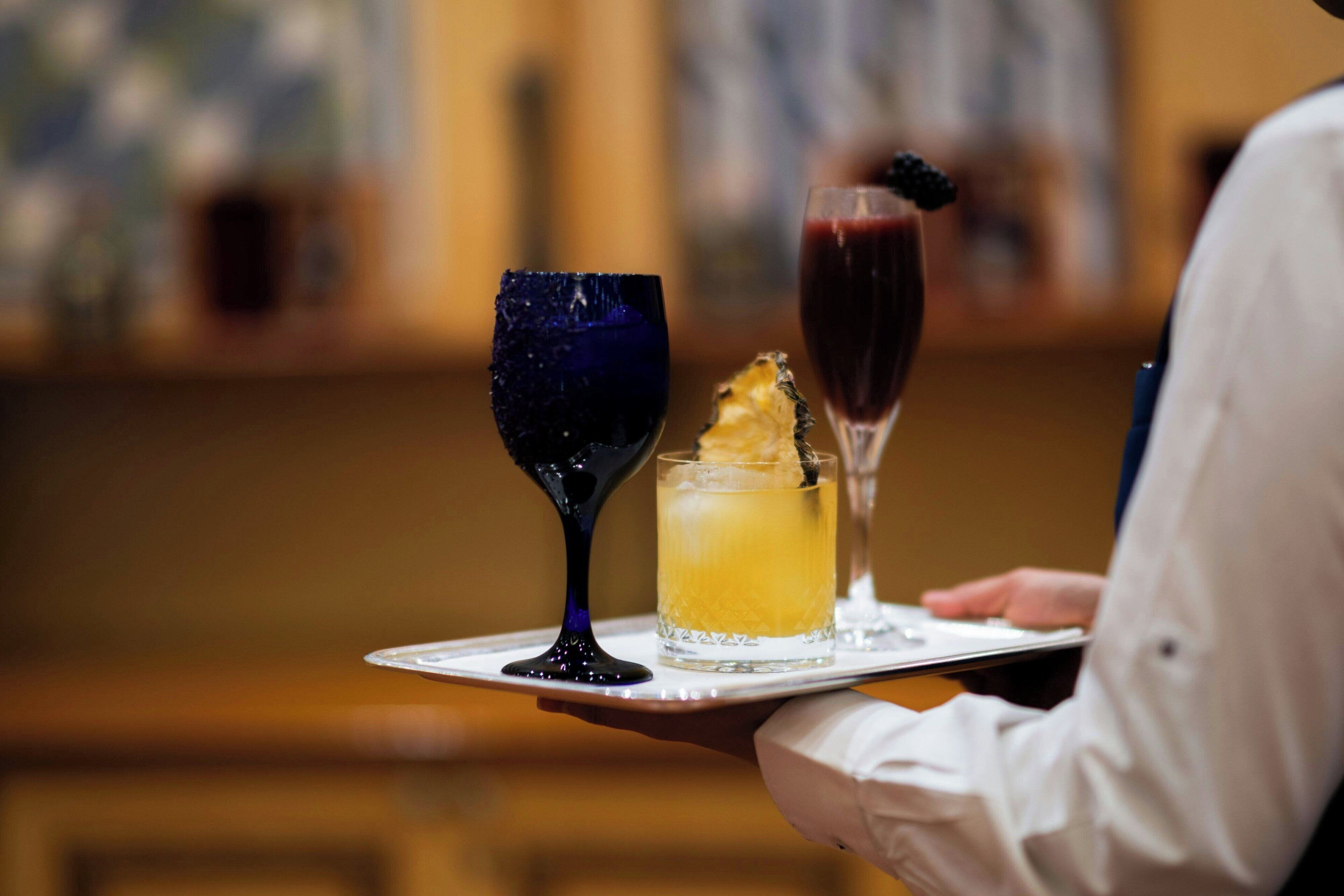Una persona sostiene una bandeja con tres bebidas, una en un vaso azul, una en un vaso amarillo y una en un vaso rojo con una baya negra en el borde.