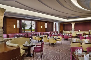 A spacious hotel dining area with multiple seating options including booths and tables, large windows offering views, and a decorative ceiling.