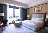 A hotel room with a large bed, two armchairs, a small table, and a view of a building through a large window.