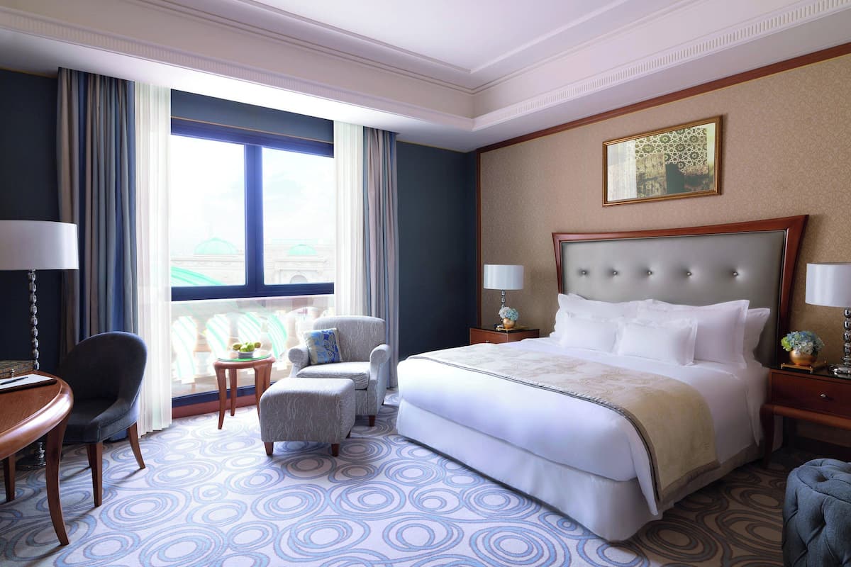Quarto de hotel com uma cama grande, duas poltronas, uma mesa pequena e vista para um edifício através de uma janela ampla.