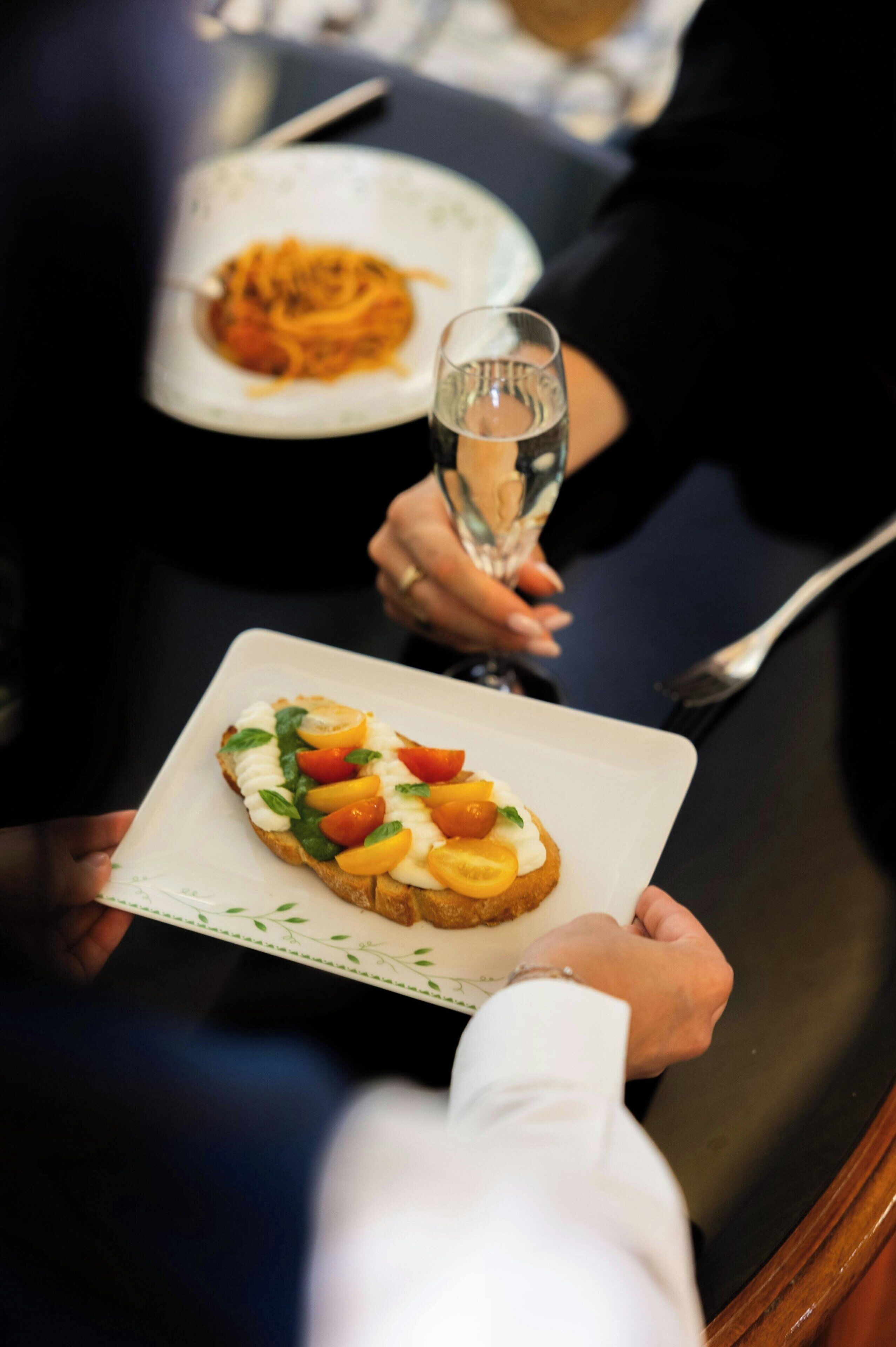 A person holding a plate with a food item topped with tomatoes and basil, and a glass of water.