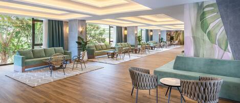 A spacious lobby with a modern design, featuring a large seating area with sofas and chairs, a wooden floor, and large windows offering a view of greenery outside.