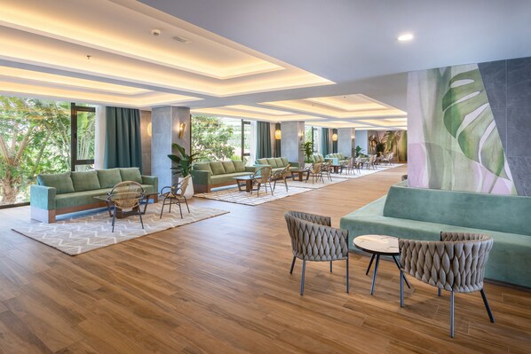 A spacious lobby with a modern design, featuring a large seating area with sofas and chairs, a wooden floor, and large windows offering a view of greenery outside.