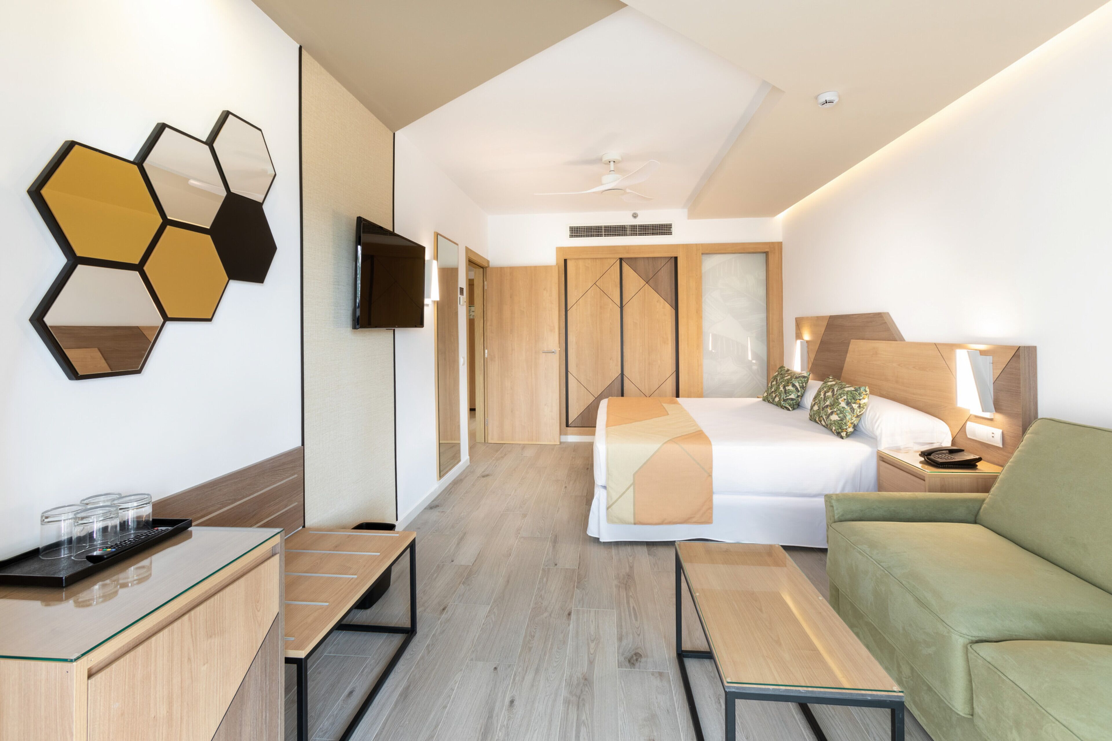 A modern hotel room with a bed, a green sofa, a desk with a glass top, and a wall-mounted TV.