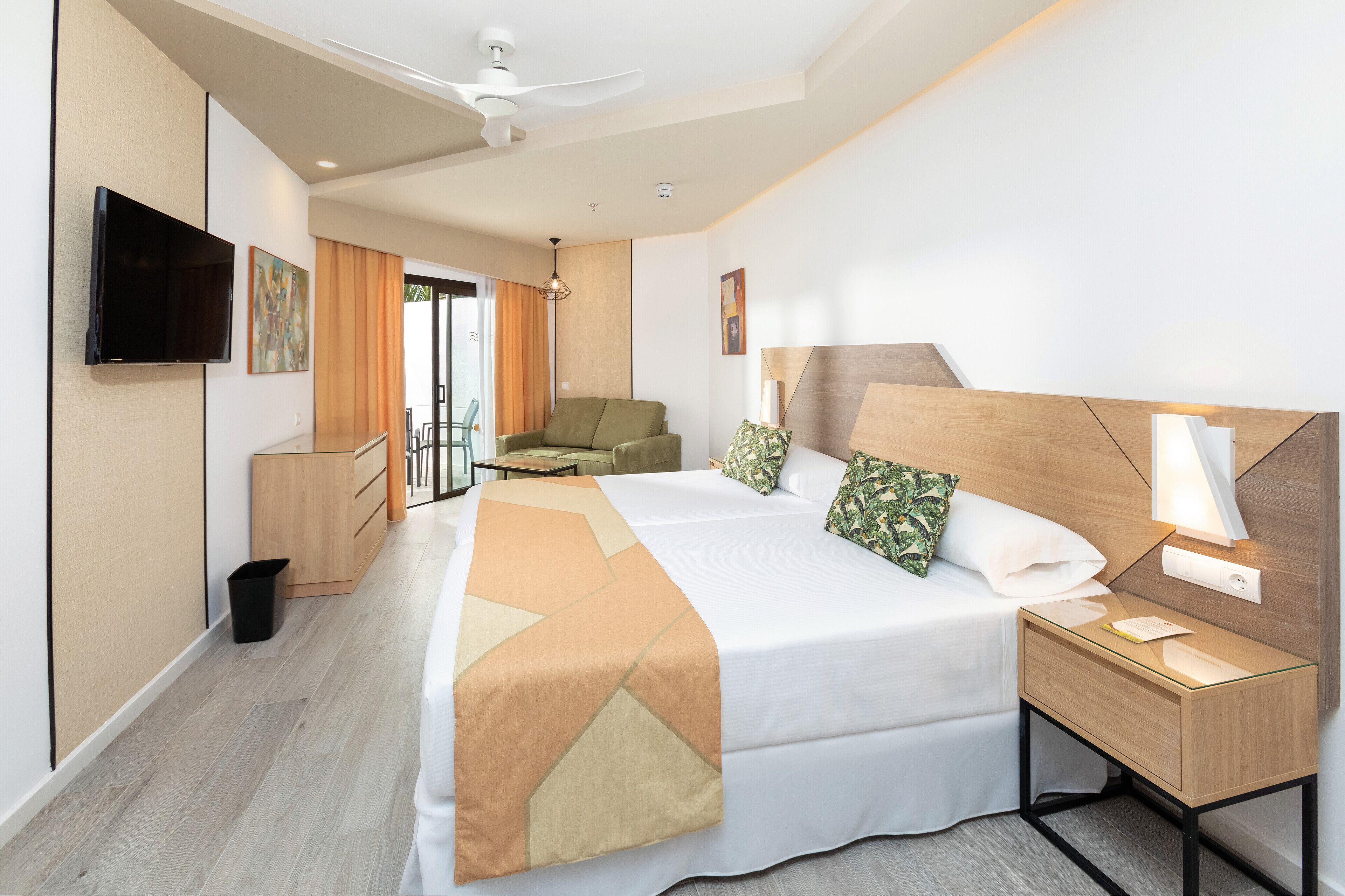 Quarto de hotel moderno com uma cama grande, mesa de cabeceira com abajur, televisão na parede e ventilador de teto.