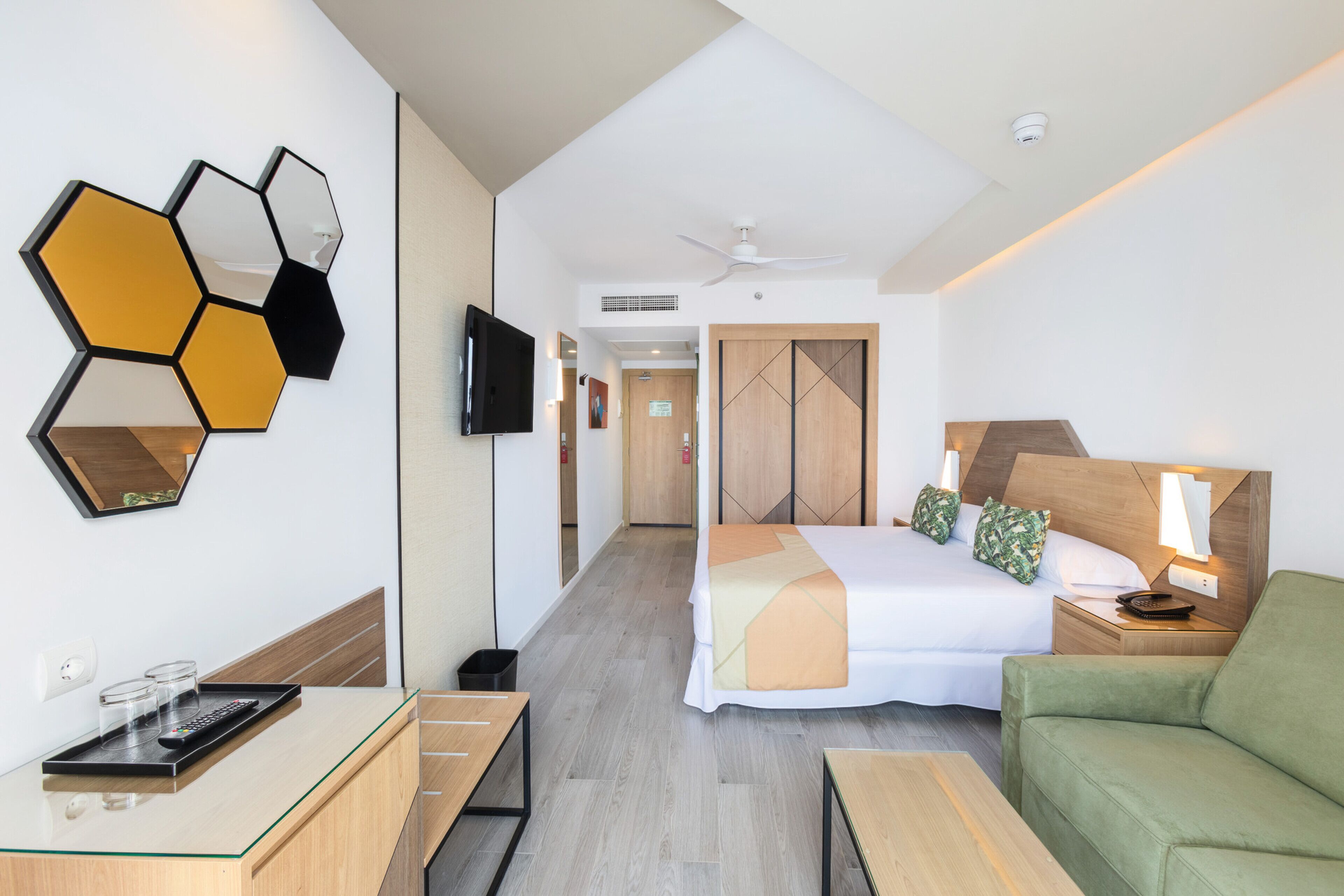 Una habitación de hotel con cama, televisión, escritorio, sofá verde y pared con espejos hexagonales.