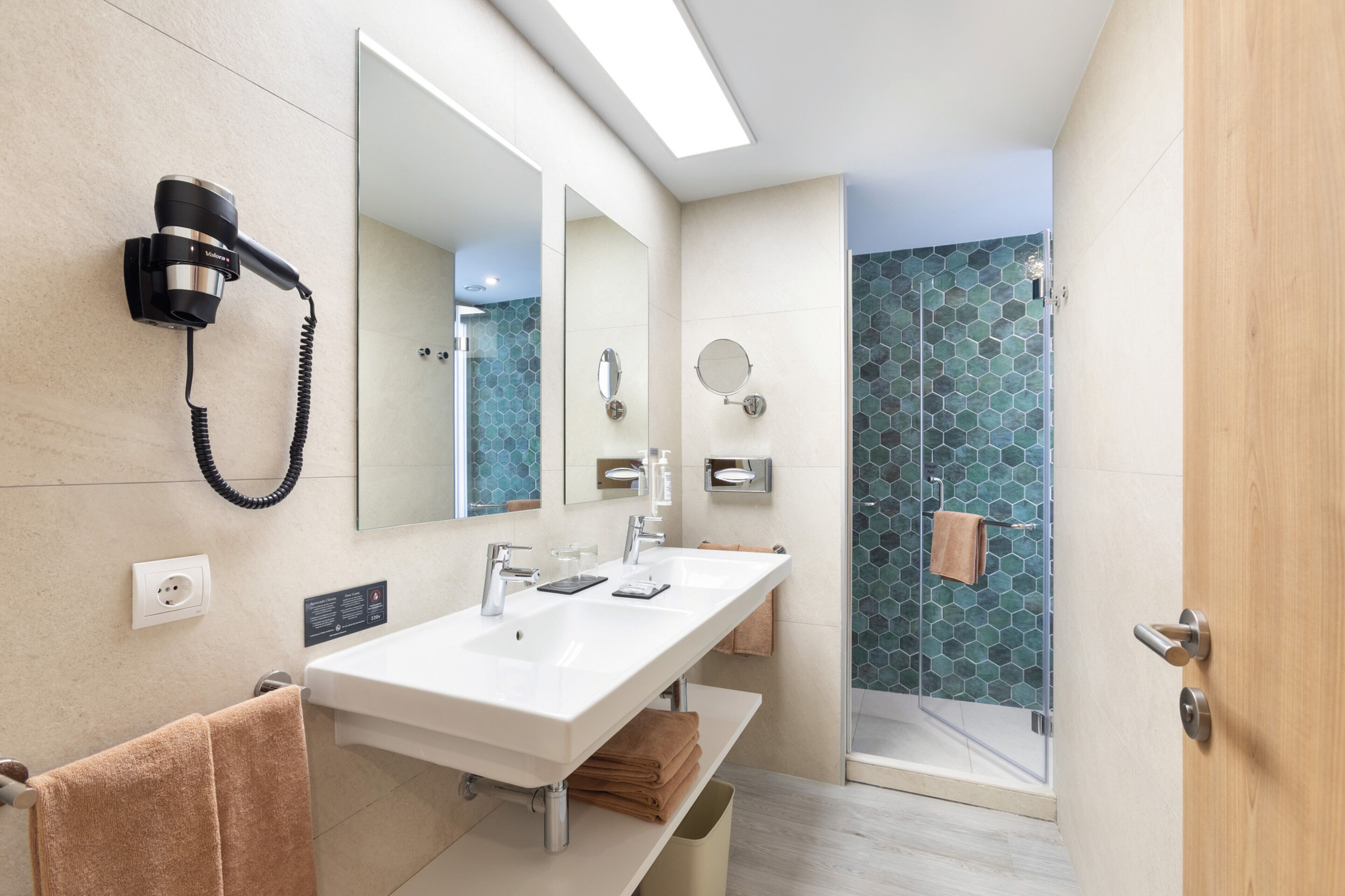 Un baño moderno con un lavabo blanco, dos espejos, un secador de pelo montado en la pared y una ducha con puerta de vidrio.