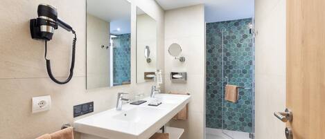 Un baño moderno con un lavabo blanco, dos espejos, un secador de pelo montado en la pared y una ducha con puerta de vidrio.