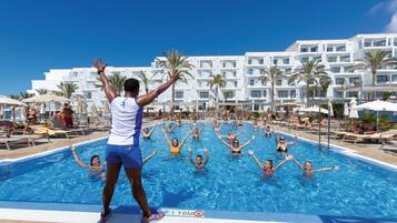 Zona de piscina con gente disfrutando del agua, rodeada por un edificio blanco con varios balcones y palmeras.
