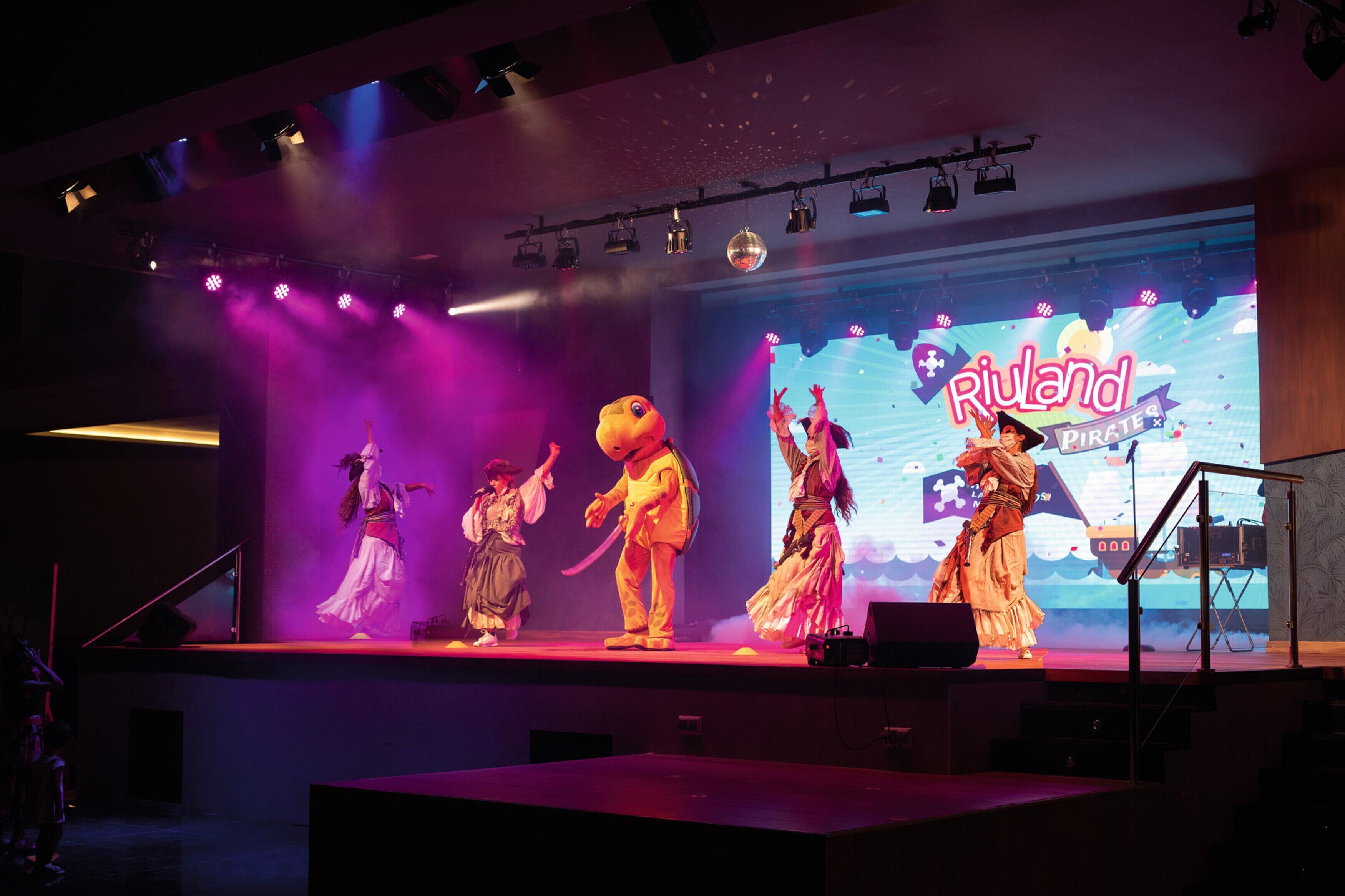 A stage with performers in costumes, a large screen displaying 'Riuland Pirate' text, and stage lighting.