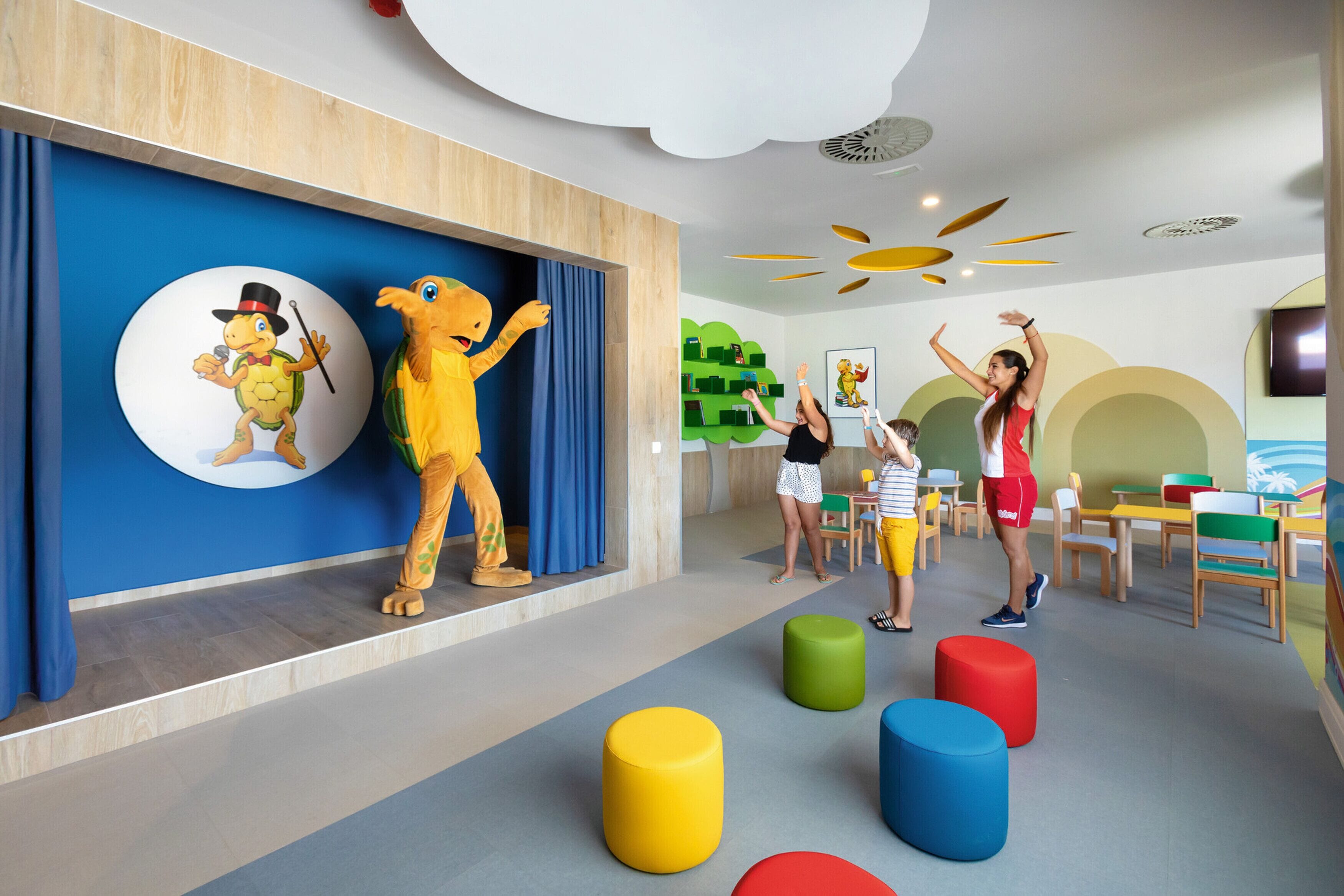 Zona de juegos infantiles con taburetes de colores, una gran mascota de tortuga y un niño jugando.