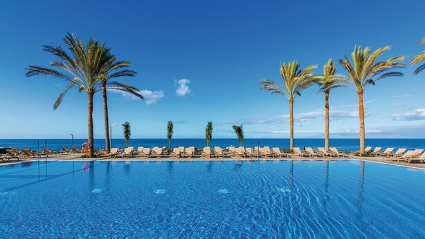 A clear blue pool with lounge chairs and palm trees, overlooking the ocean. - Hotel Riu Buenavista - All Inclusive (Adeje)