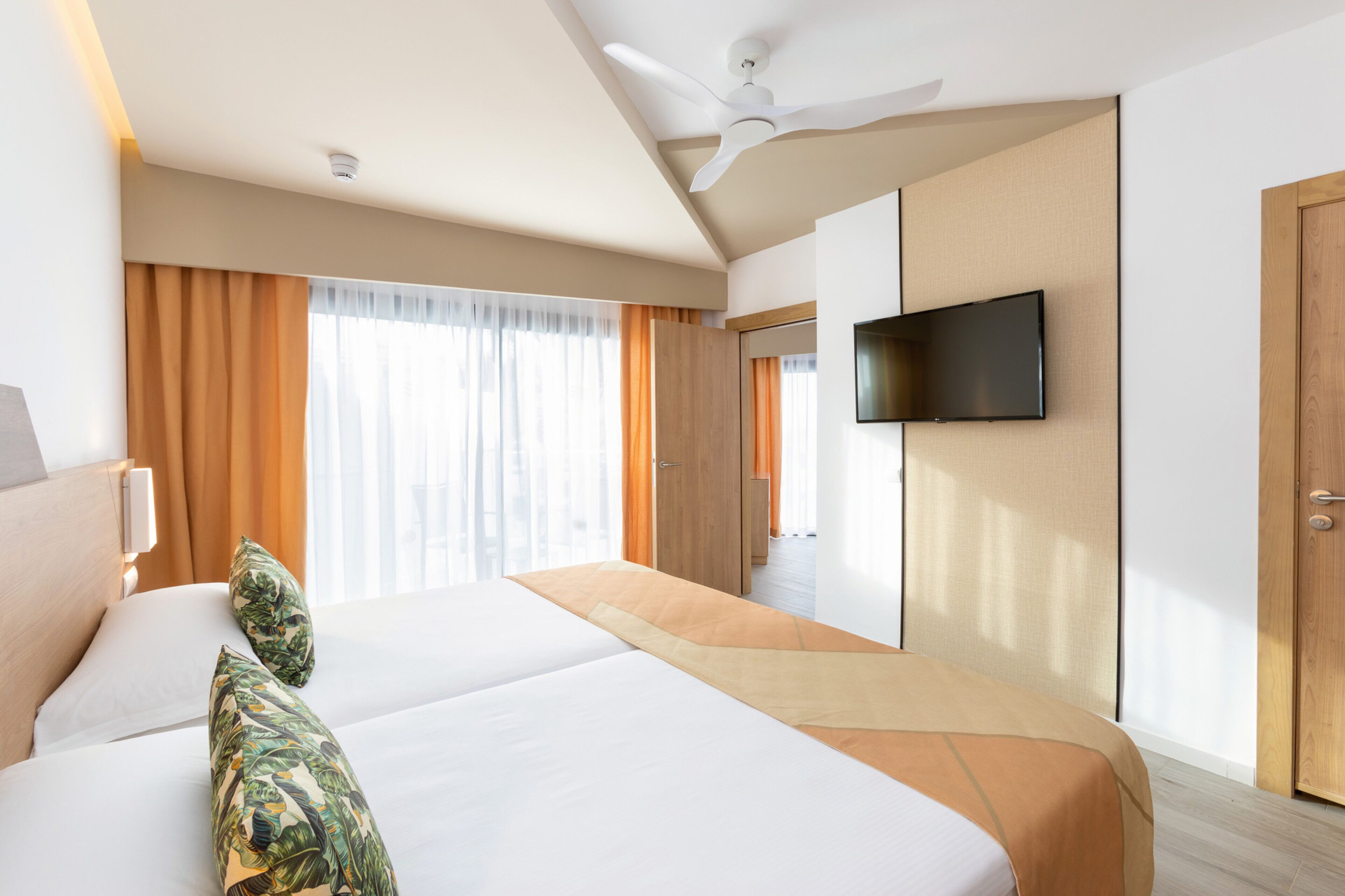 Habitación de hotel con dos camas, un televisor de pantalla plana montado en la pared, ventilador de techo y ventanas grandes con cortinas.