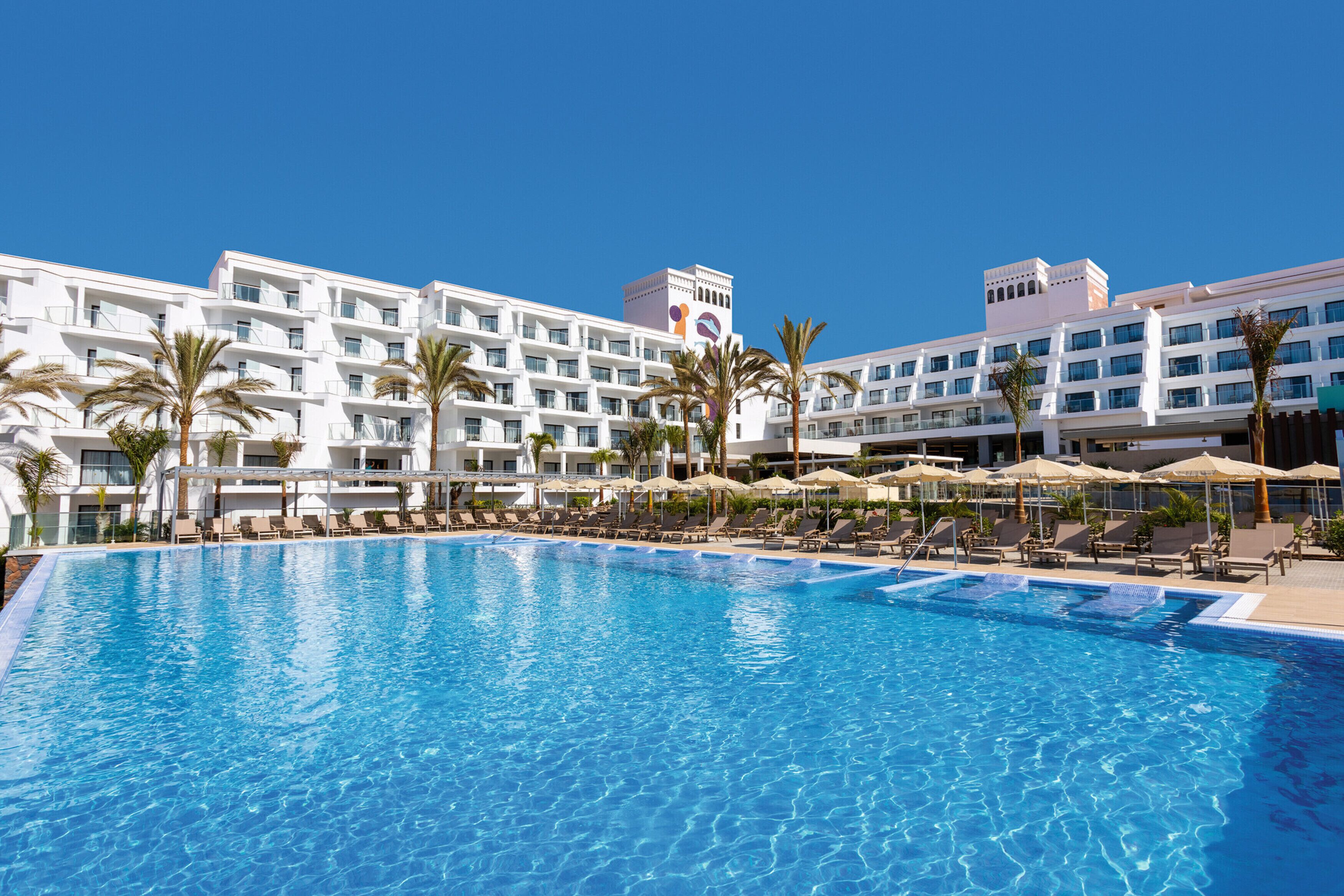 A hotel with a large swimming pool, palm trees, and multiple balconies.