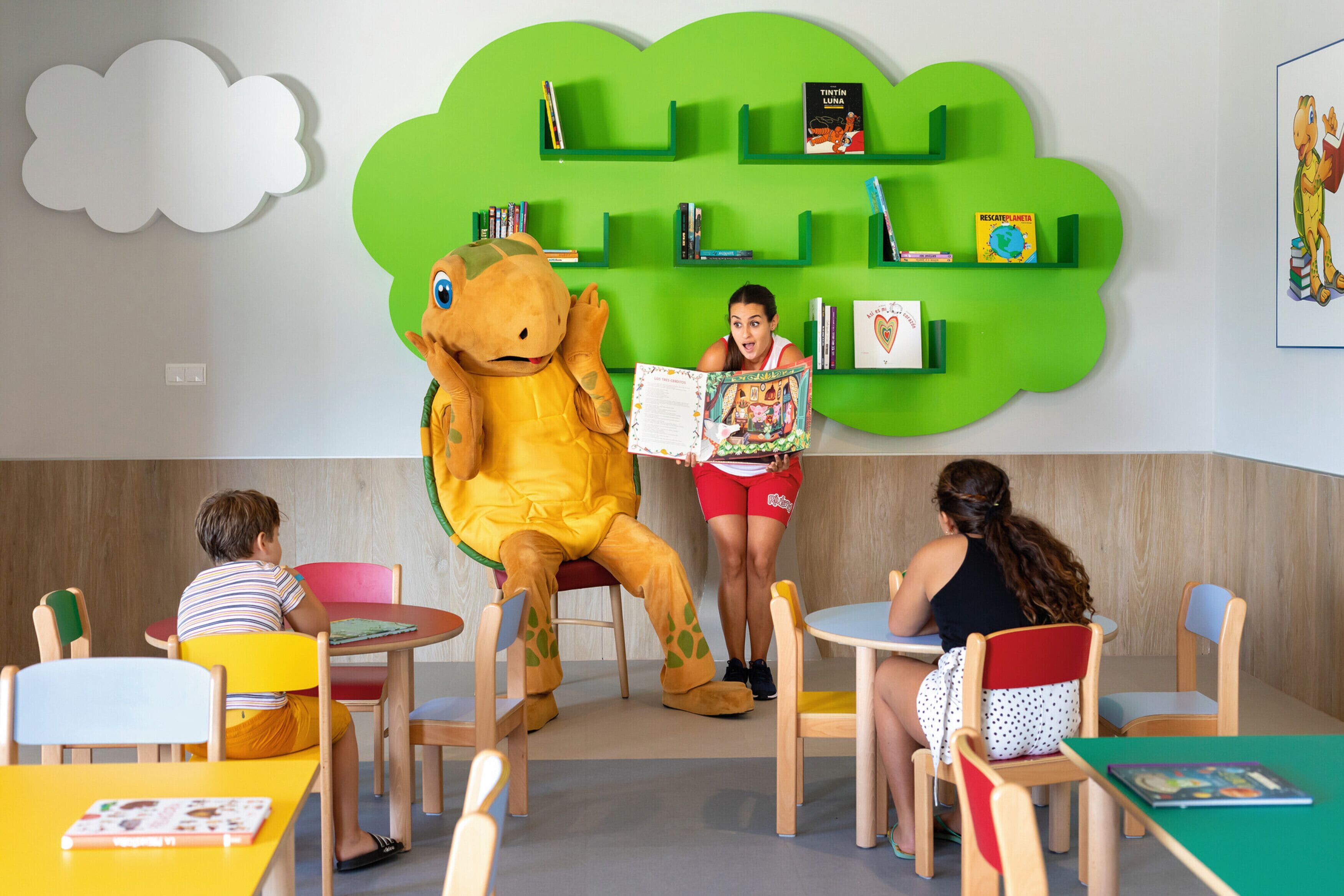 Una biblioteca infantil con una estantería verde, una mascota de tortuga y niños leyendo.