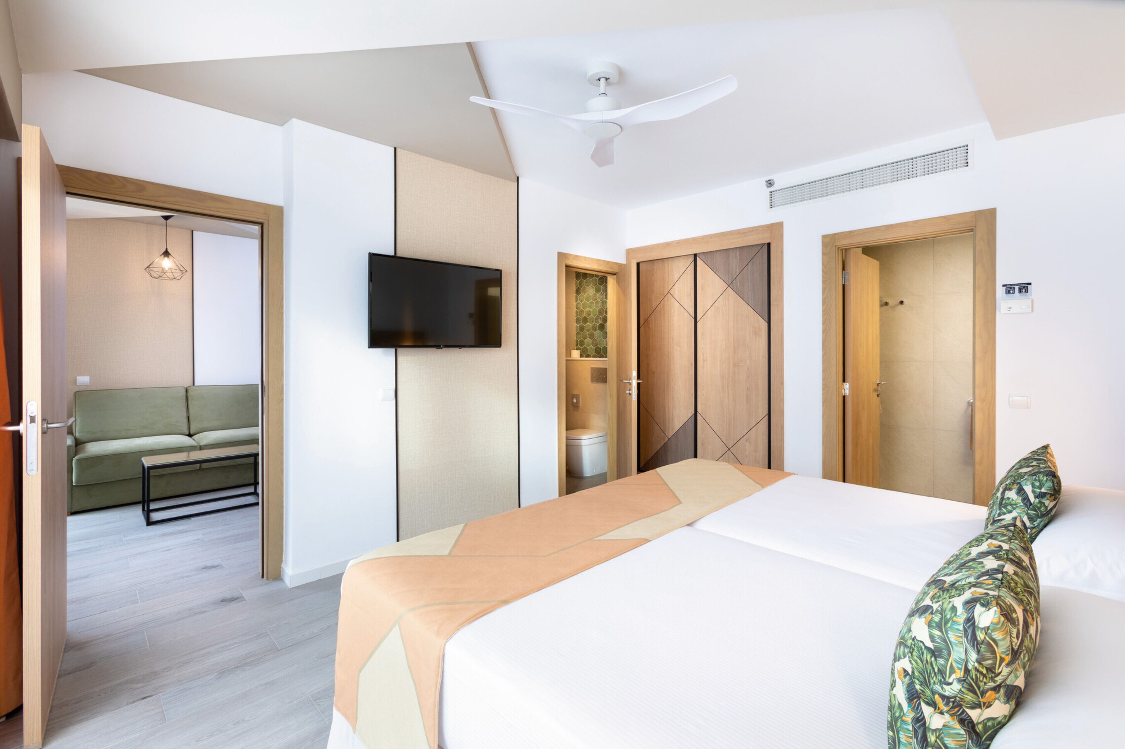 Ett modernt hotellrum med en säng, en TV, en takfläkt och utsikt över ett annat rum med en soffa och en hängande lampa.