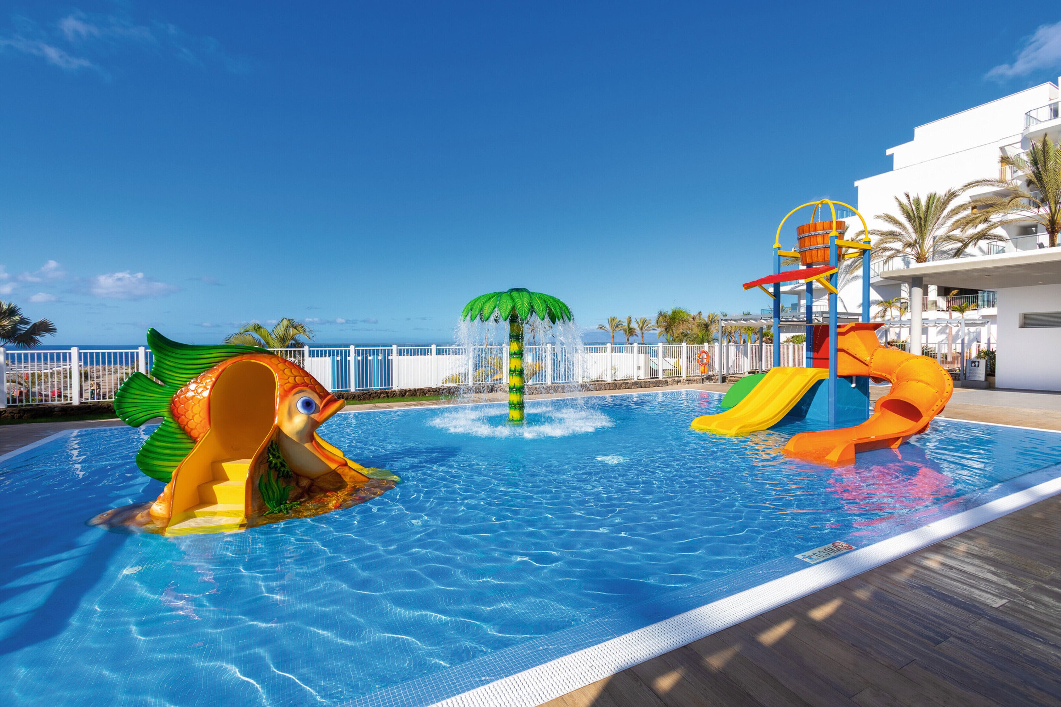 A swimming pool with a fish-shaped slide and a water play area with a slide and a bucket lift.