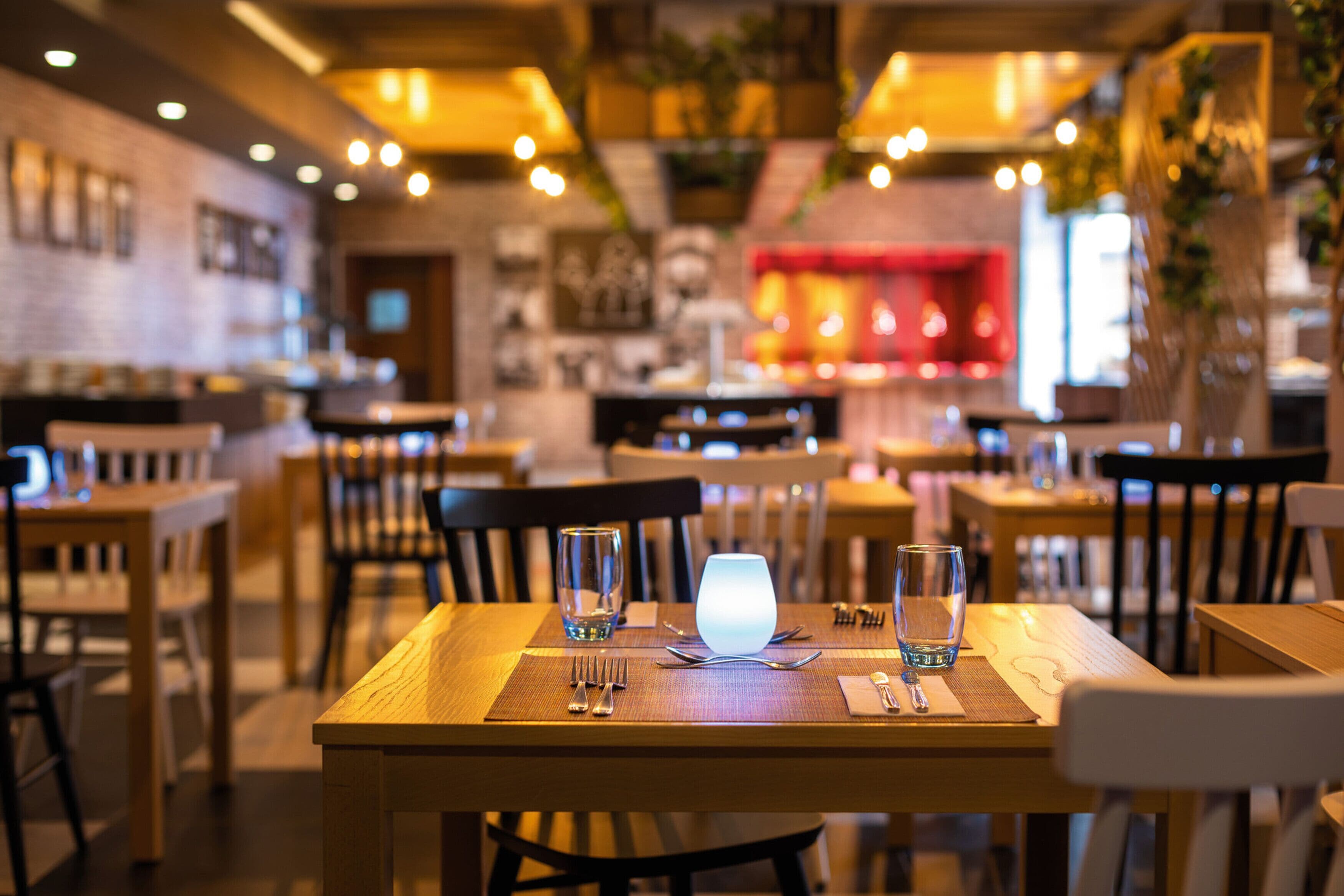A restaurant with wooden tables and chairs, a lit table lamp, and a bar area in the background.