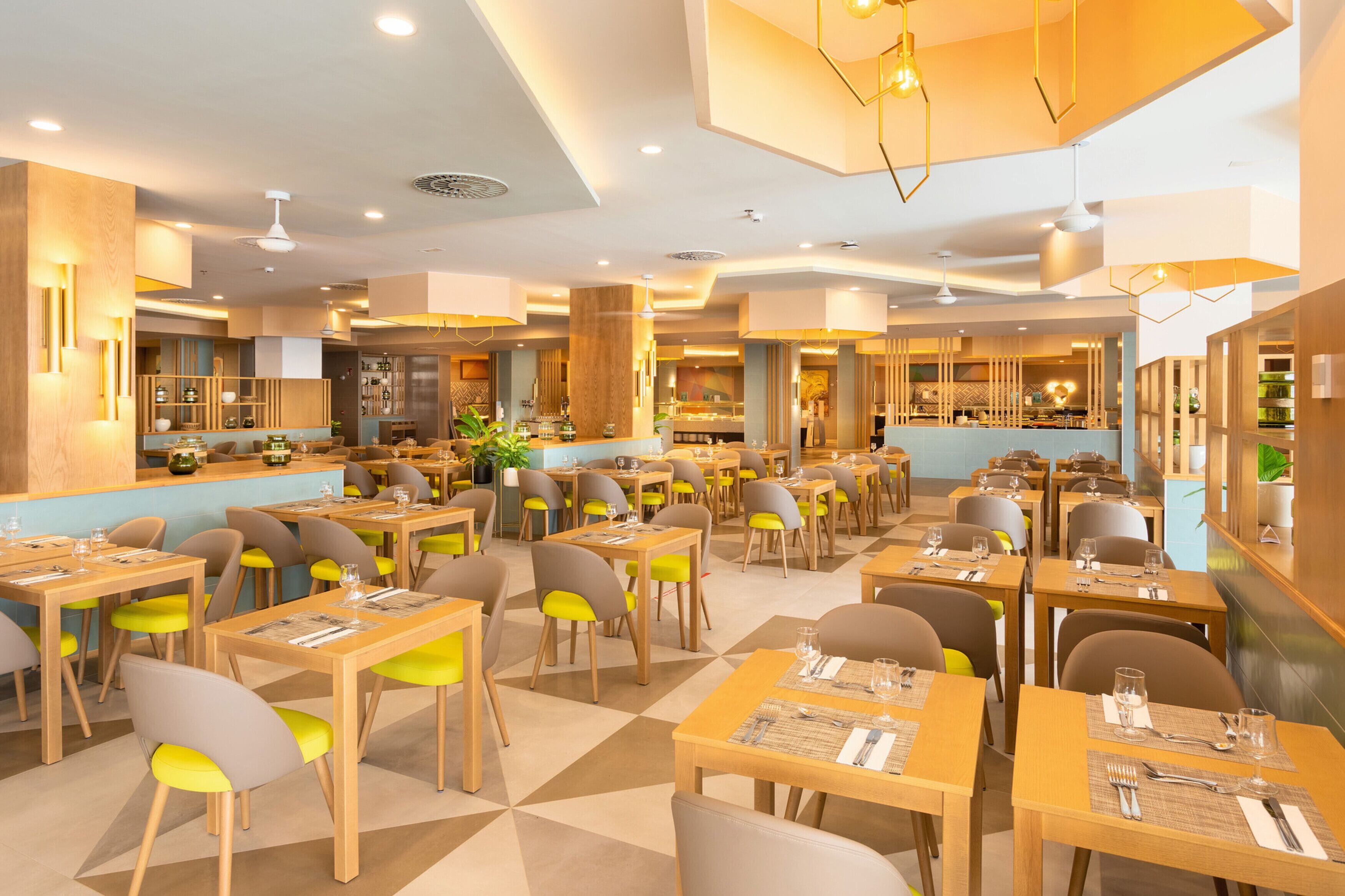 A spacious restaurant with multiple dining tables and chairs, a kitchen area in the background, and modern lighting fixtures.