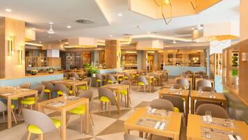 A spacious restaurant with multiple dining tables and chairs, a kitchen area in the background, and modern lighting fixtures.