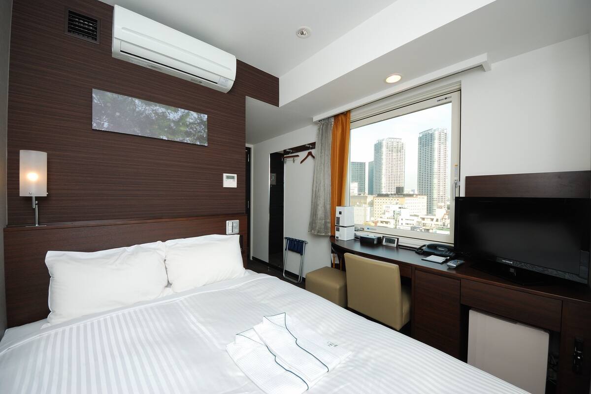 A hotel room with a bed, a desk, a television, and a window with a city view.