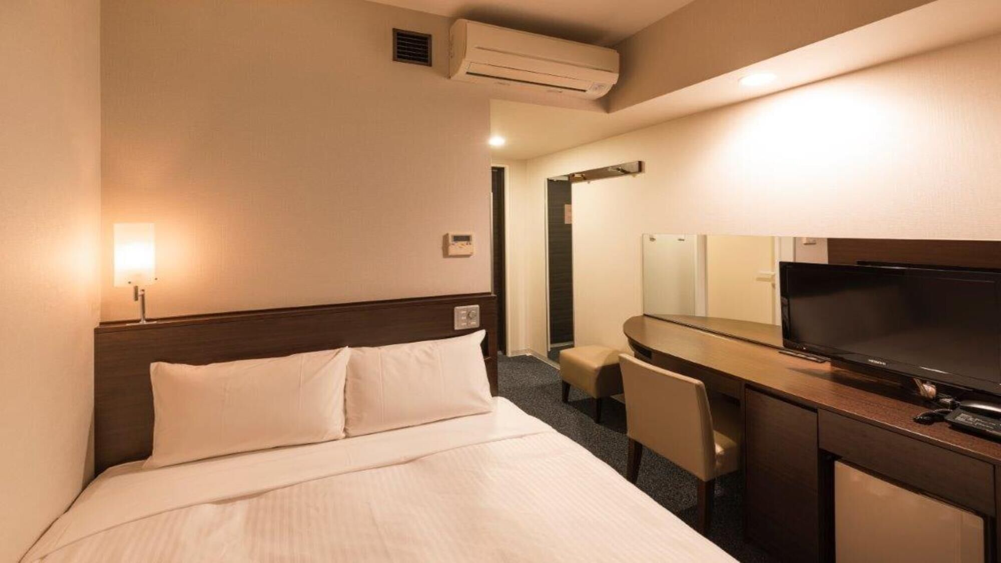 Een hotelkamer met een bed, een bureau met een televisie, een stoel en een airconditioning.