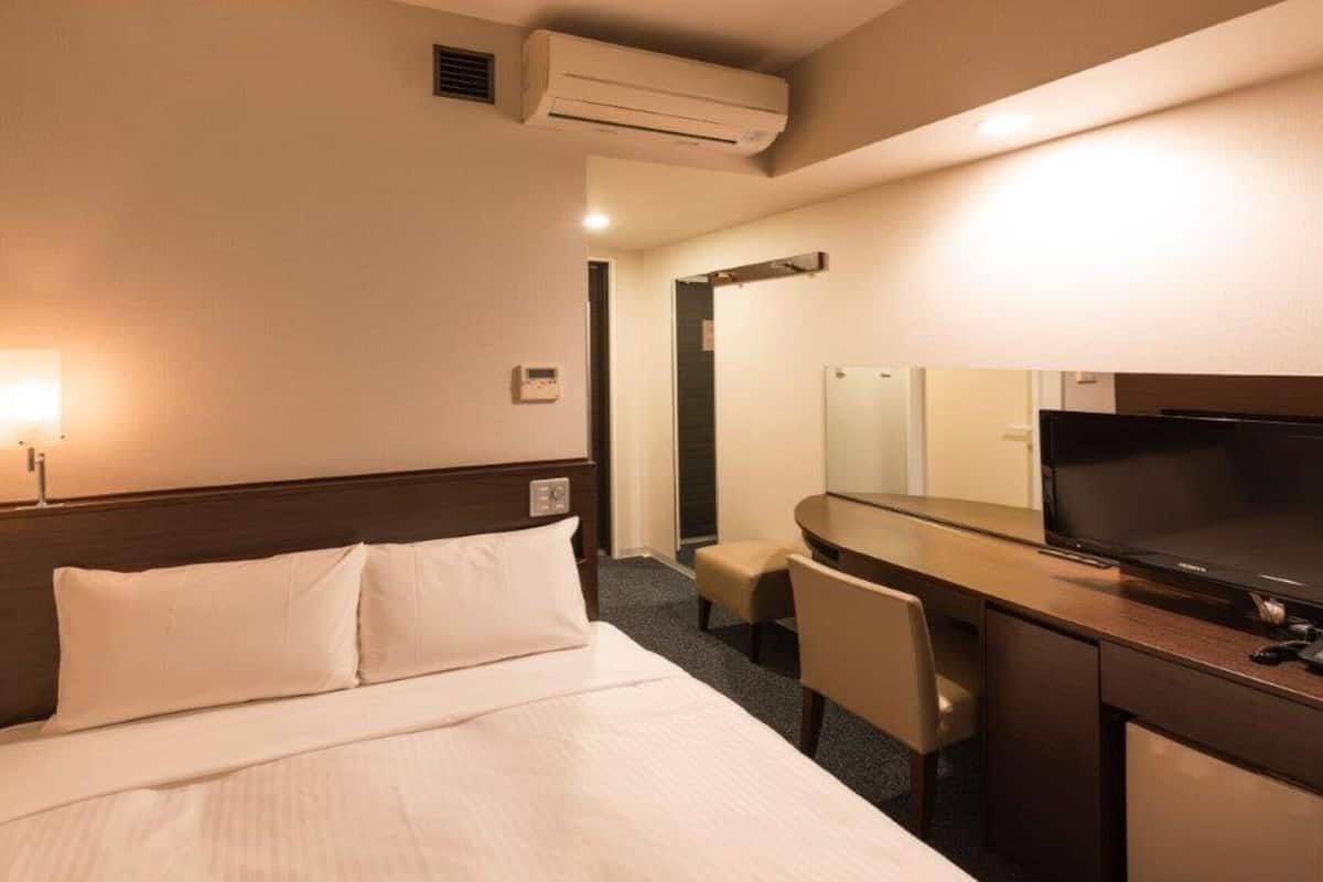 A hotel room with a bed, a desk with a TV, a chair, and an air conditioning unit.