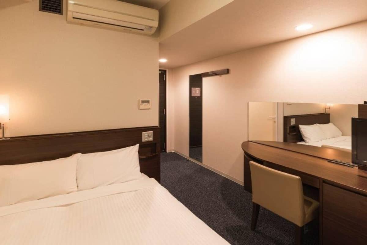 Quarto de hotel com cama, escrivaninha, cadeira, televisão e espelho.