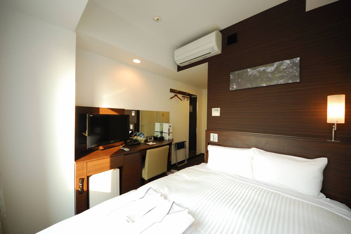 A hotel room with a bed, a desk with a TV, a mirror, and a wall-mounted air conditioner.
