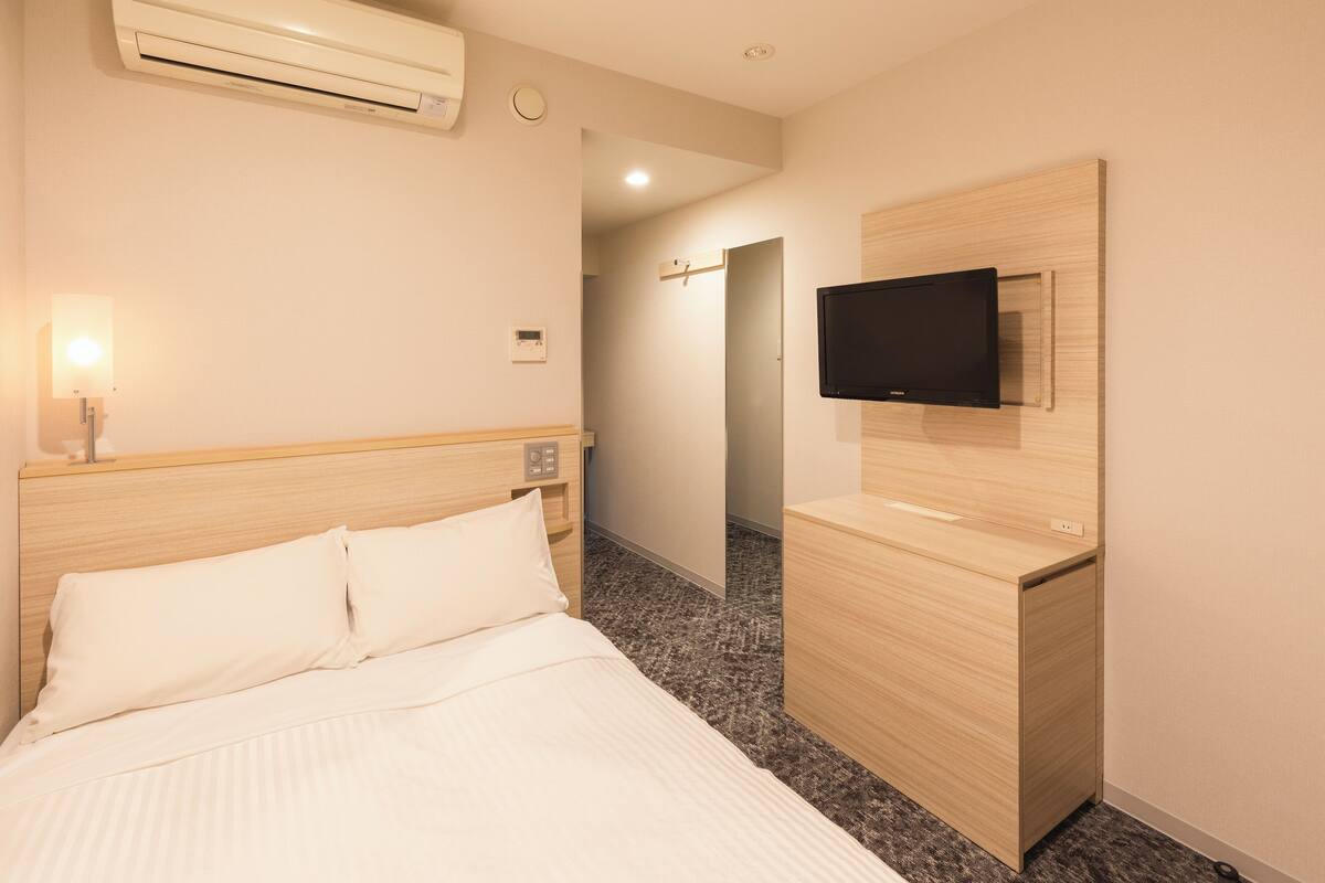 A hotel room with a bed, a TV mounted on a wooden cabinet, and an air conditioning unit on the wall.