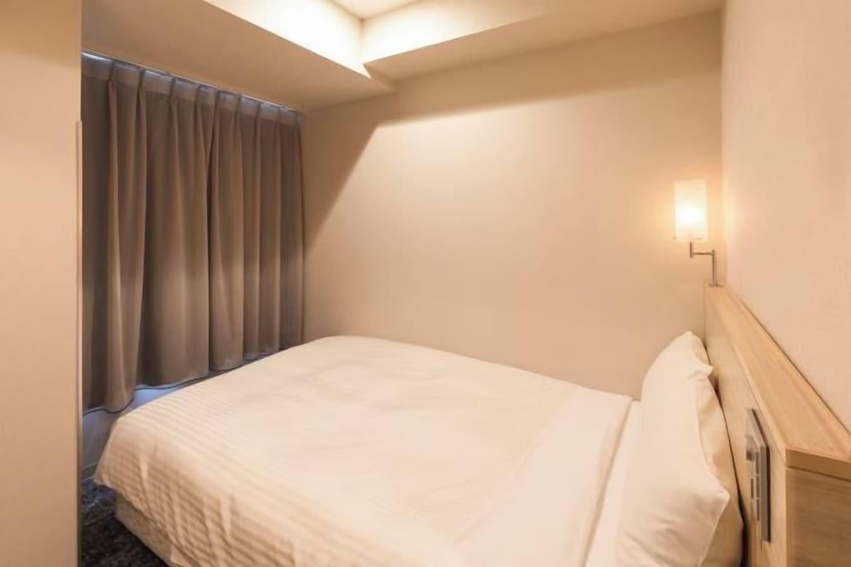 A hotel room with a bed, a nightstand, a wall-mounted lamp, and a window with curtains.