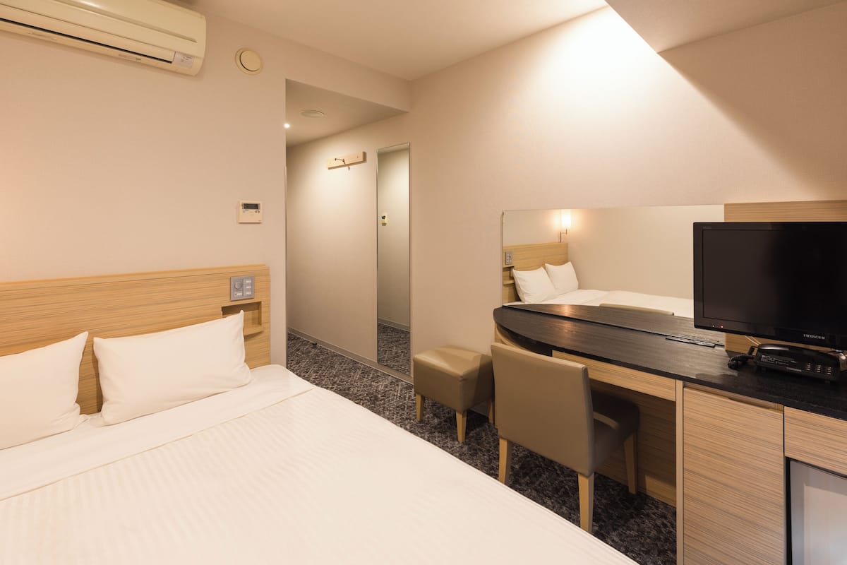A hotel room with a bed, a desk with a chair, a television, and an air conditioning unit.