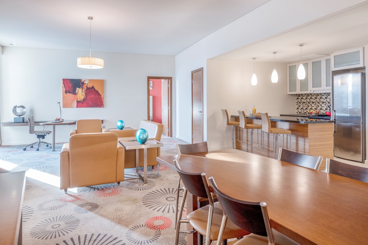 A modern kitchen with a stainless steel refrigerator, a bar area with stools, and a dining area with a large wooden table and chairs.