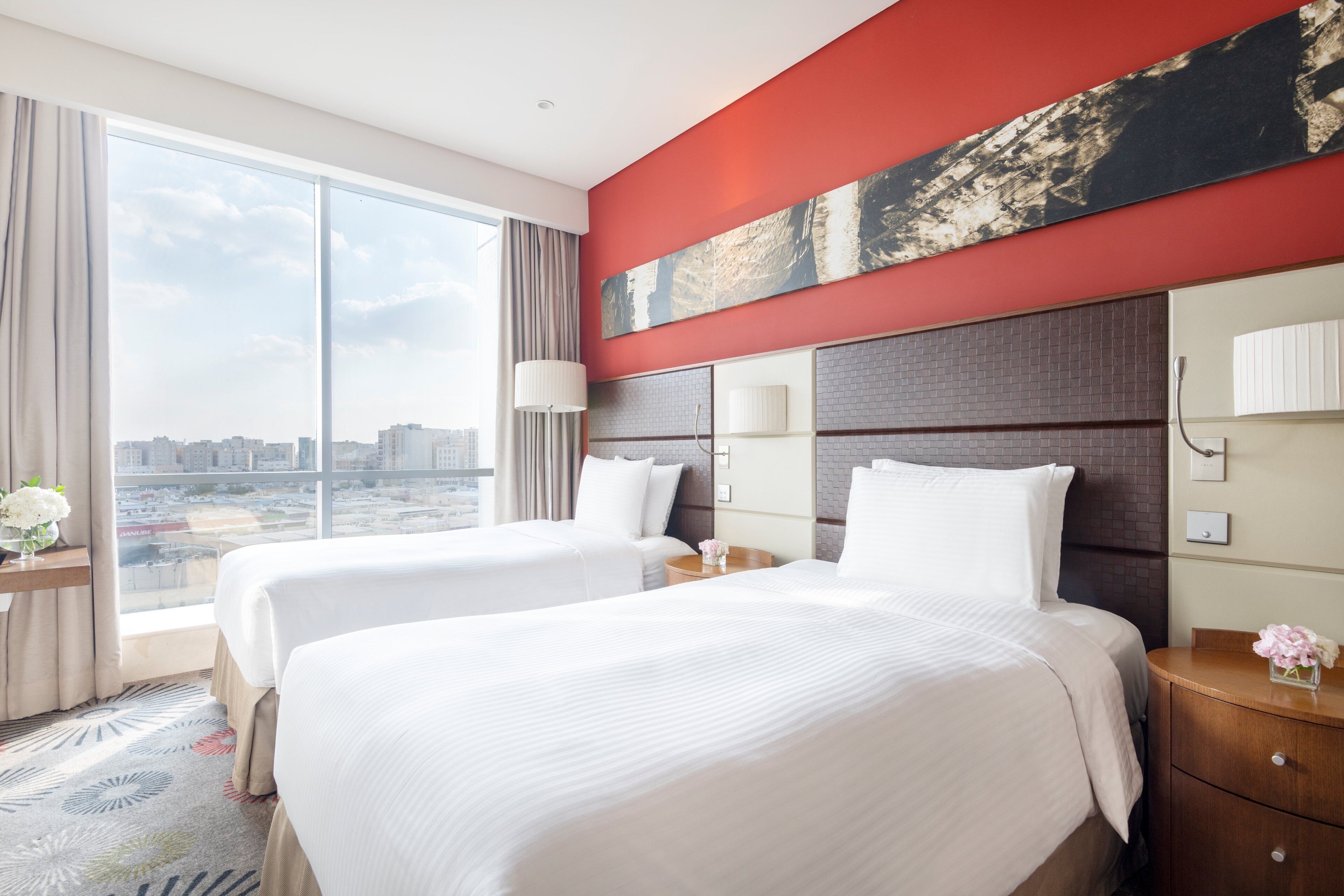 A hotel room with two beds, a large window with a city view, a red accent wall, and a modern design.