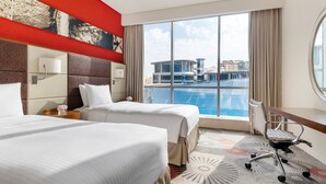 A hotel room with two beds, a desk, a chair, a large window with a city view, and a red accent wall.