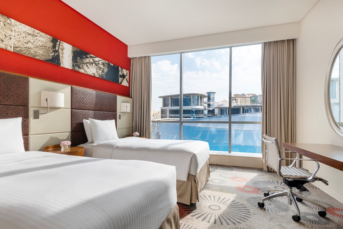 A hotel room with two beds, a desk, a chair, a large window with a city view, and a red accent wall.