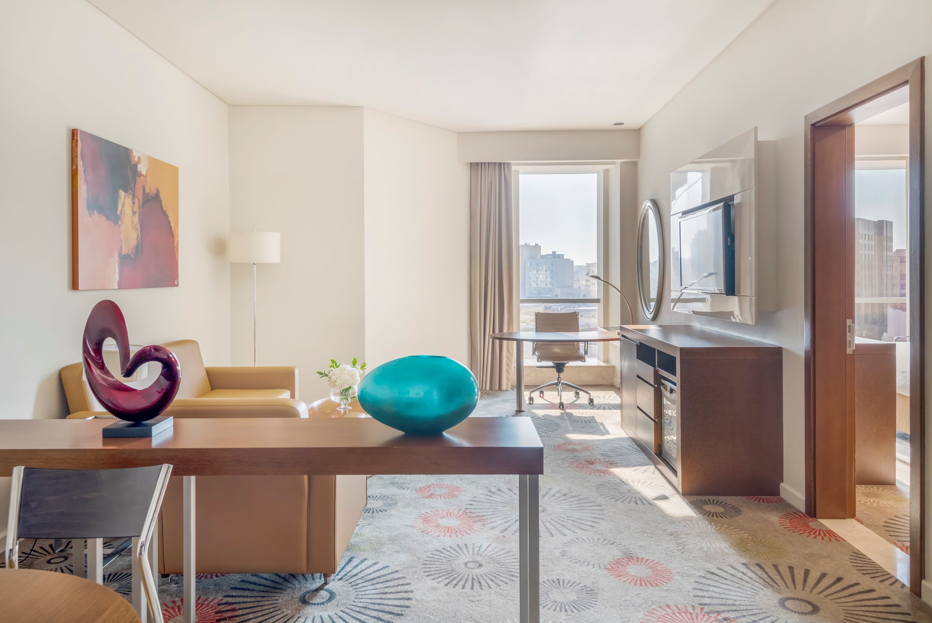 A hotel room with a dining area, a sofa, a TV, and a window with a view of buildings.