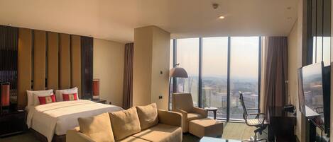 A hotel room with a bed, sofa, armchair, desk, and large windows offering a city view.