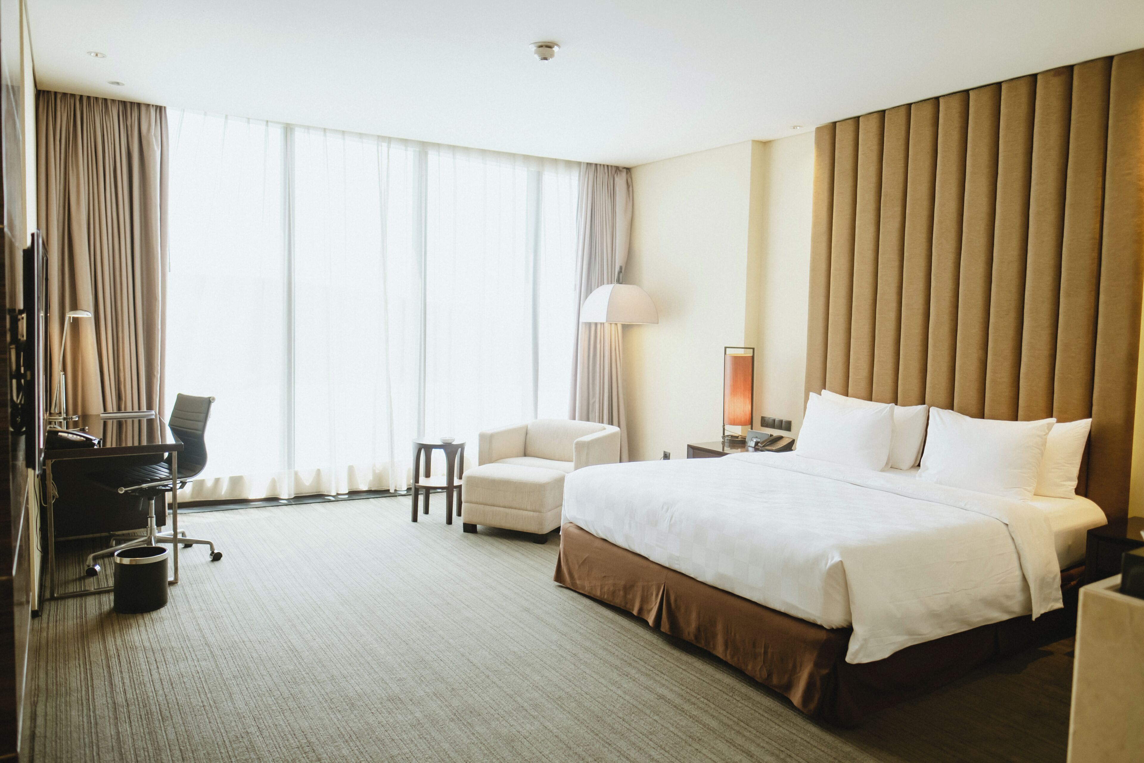 A hotel room with a large bed, a desk with a chair, a small armchair, a lamp, and a window with curtains.