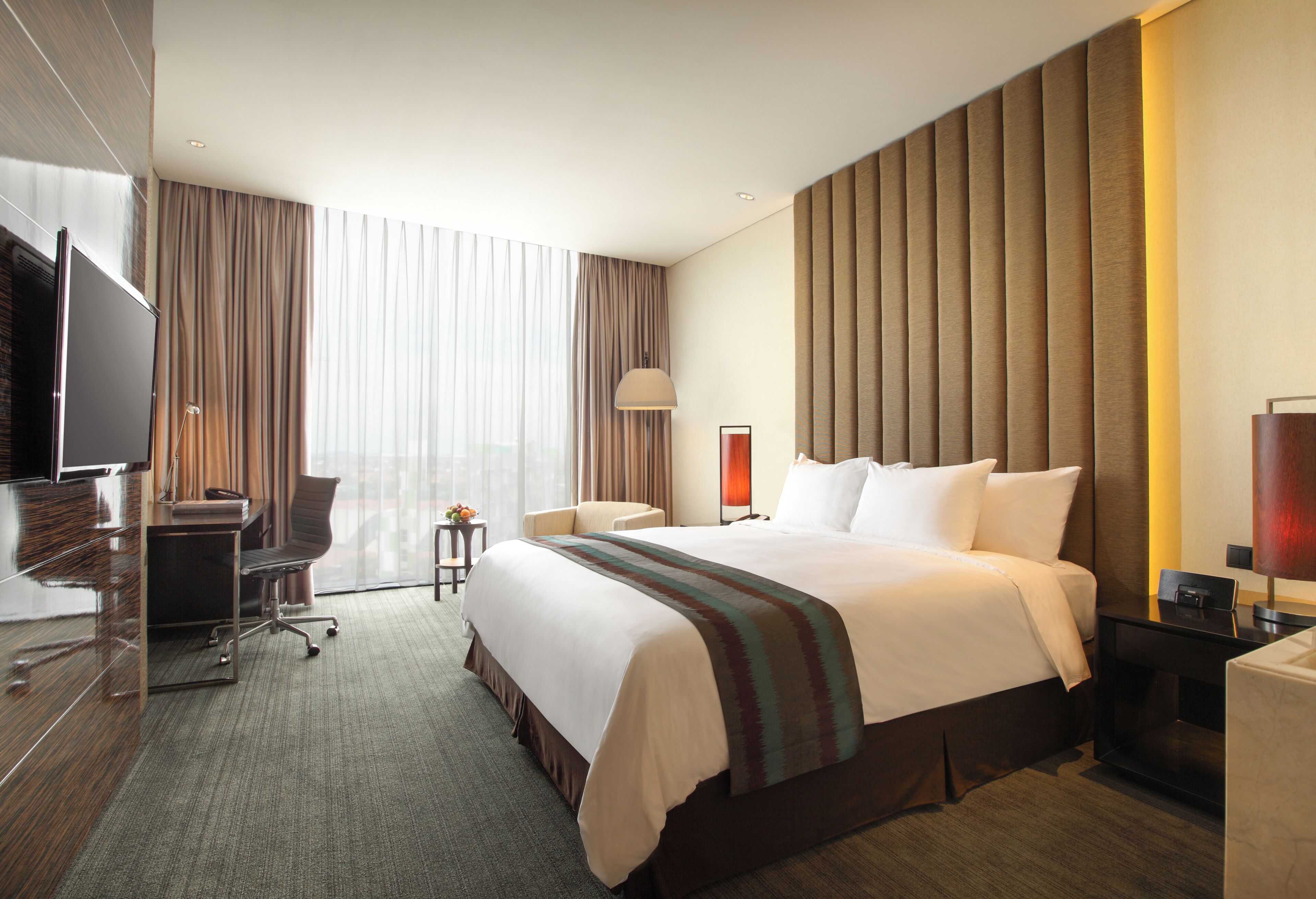 A hotel room with a bed, desk, chair, TV, and a large window with curtains.