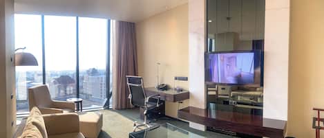 A modern hotel room with a large window, a sofa, a glass coffee table, a desk, and a flat-screen TV.