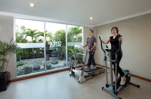 A room with a treadmill, elliptical, and stationary bike, large windows with a view of a garden, and a potted plant.