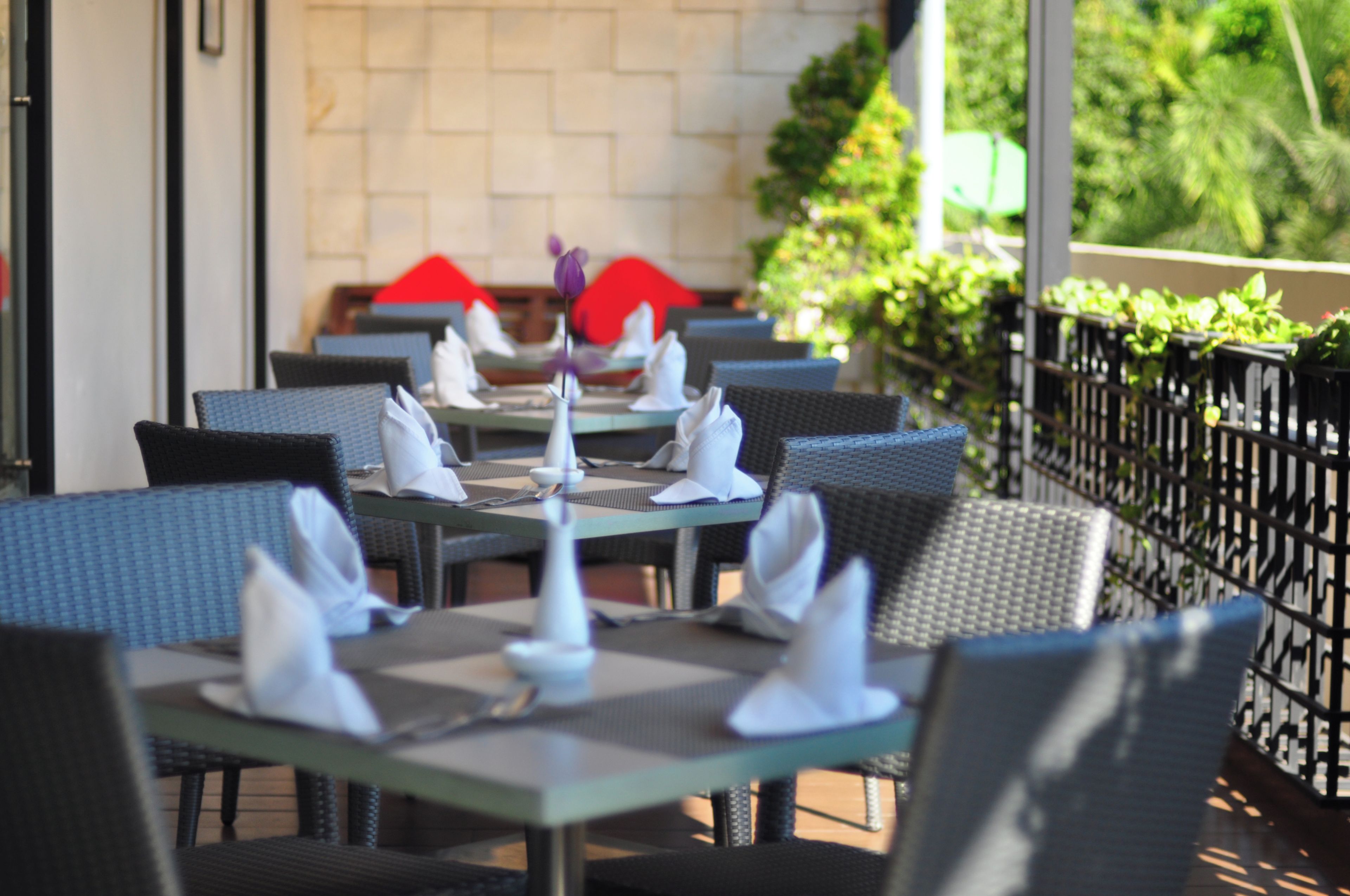 An outdoor dining area with tables set for two, featuring wicker chairs and a tiled wall in the background.