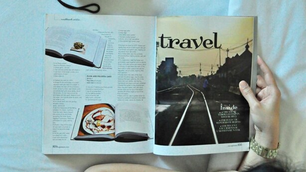 A person lying on a bed with a book open to a travel magazine, a camera, and a smartphone. - Hotel Santika Jemursari Surabaya (Surabaya)