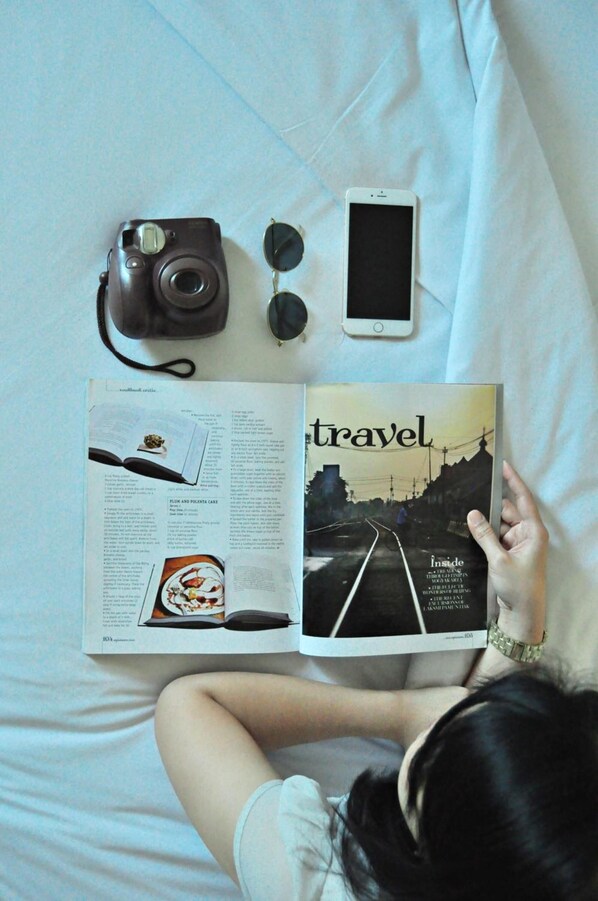 A person lying on a bed with a book open to a travel magazine, a camera, and a smartphone. - Hotel Santika Jemursari Surabaya (Surabaya)