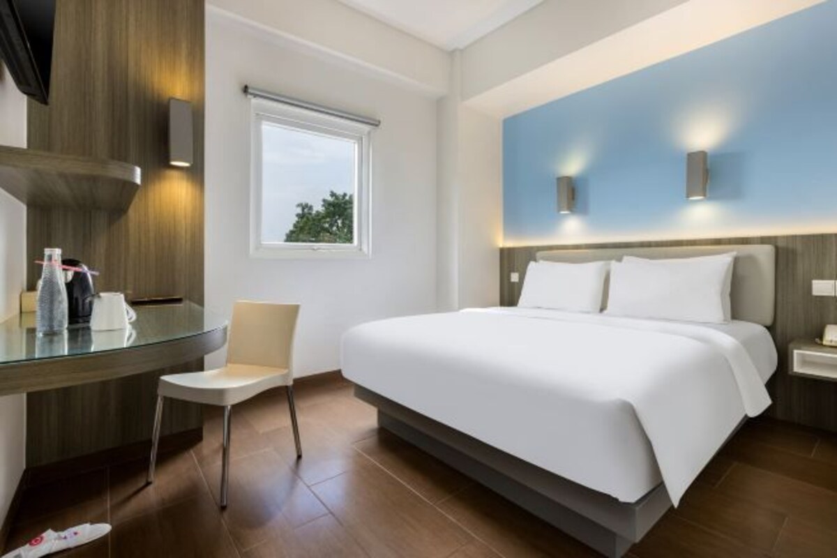 Une chambre d’hôtel moderne équipée d’un lit, d’un bureau avec une chaise, d’une fenêtre et d’un luminaire fixé au mur.