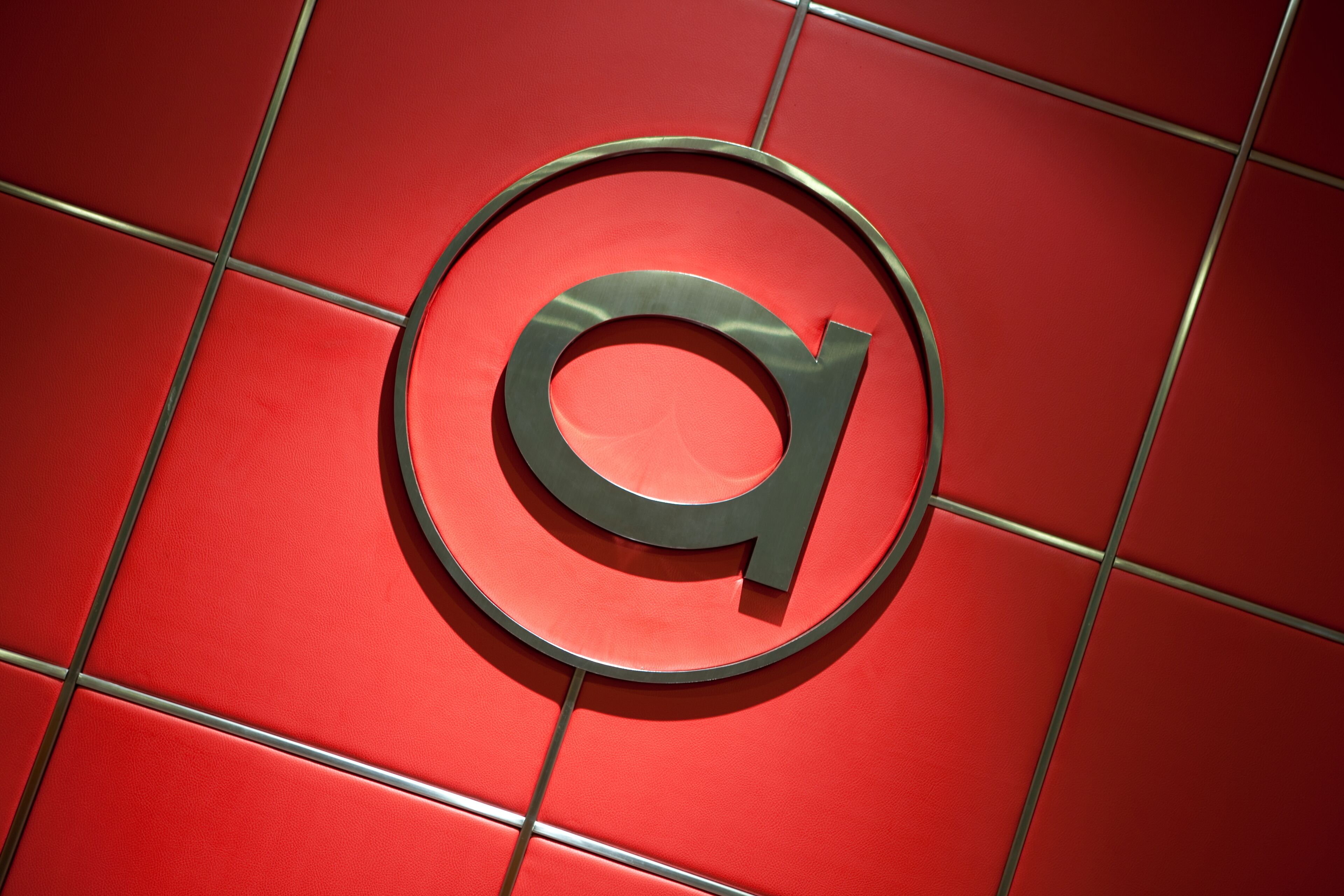 Un logotipo metálico y circular con una 'a' minúscula sobre un muro de baldosas rojas.